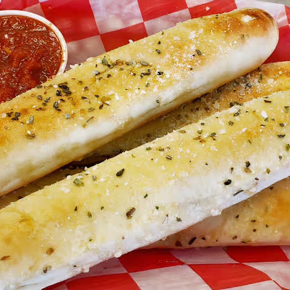 Three herb-seasoned toasted breadsticks served with marinara sauce on red-and-white checkered paper.